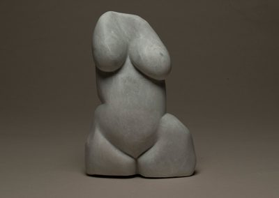 A stone sculpture of a womans body with rolls and uneven breasts and thick thighs.