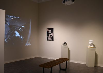 A video playing on a wall in the memoirs exhibition next to a skeleton painting and two sculptures.